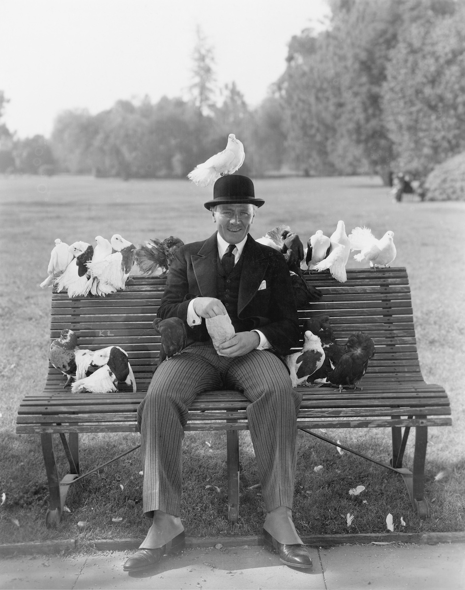 A gentleman in a bowler hat on a park bench, feeding pigeons